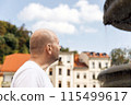 Bald man in white t-shirt looking up, city buildings in background, blue sky 115499617