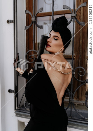 Stylish woman in the city. Fashion photo of a beautiful model in an elegant black dress posing against the backdrop of a building on a city street Stylish woman in the city. Fashion photo of a beautiful model in an elegant black dress posing against the backdrop of a building on a city street 115500213