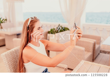 A woman is sitting at a table with a cell phone in her hand. She is taking a picture of the ocean. 115500228
