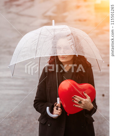 Sad middle-aged woman in a black coat and boots under a transparent umbrella. He holds a balloon in the form of a heart in his hands. Rain in the city, outdoor scene, water drops on the umbrella. The 115501265