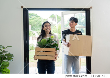 Happy Couple Moving into New House, Carrying Boxes and Plants, Fresh Start in Modern Home, Relocation and New Beginnings, Young Adults Unpacking and Settling in Bright and Airy Living Space 115501410