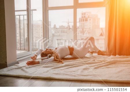 Side view portrait of relaxed woman listening to music with headphones lying on carpet at home. She is dressed in a white tracksuit. 115501554