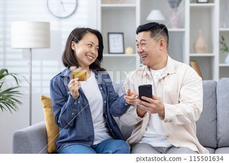 Smiling couple making an online purchase using a phone and credit card while sitting on the sofa at home. Smiling couple making an online purchase using a phone and credit card while sitting on the sofa at home. 115501634