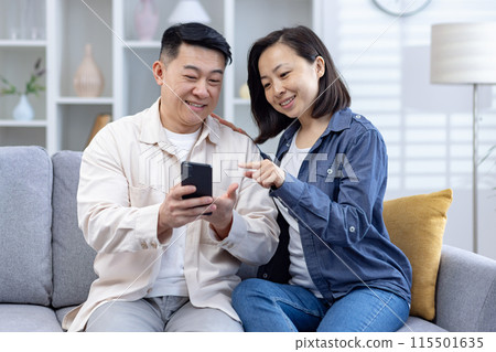Happy couple sitting on a couch and using a smartphone together in a comfortable and cozy living room. 115501635
