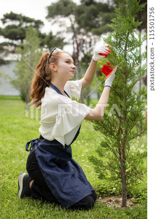 Side view of pretty female with red hair growing plants, touching tree. Concept of taking care of plants. Side view of pretty female with red hair growing plants, touching tree. Concept of taking care of plants. 115501756