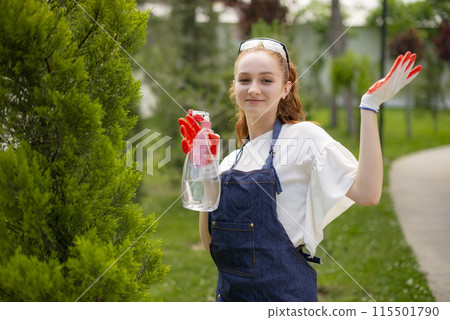 Front view of young woman spraying tree, showing, raising hand, looking at camera. Concept of taking care of plants. 115501790
