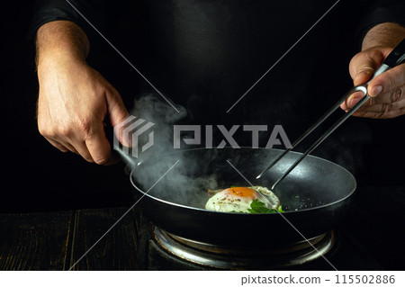 Cooking scrambled eggs in a frying pan. The chef holds a fork and a frying pan with a fried egg. Low key concept of preparing breakfast 115502886