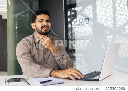 A young Indian man fell ill at work, sits at his desk, works on a laptop, holds his hand to his neck, feels a strong pain in his throat. 115504060