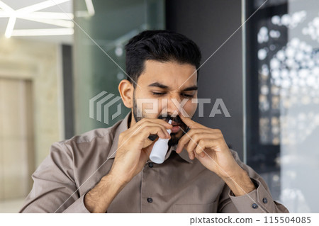 A close-up photo of a young Indian man who is being treated while in the office at the workplace, blowing his nose with medication for a sore throat. 115504085