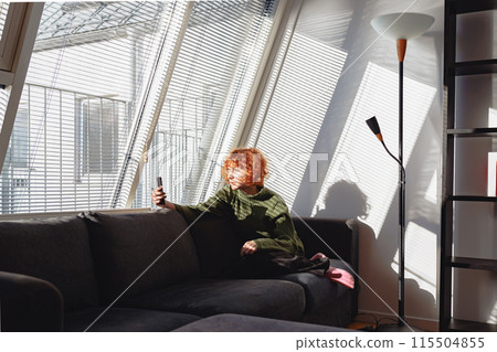 young red-haired woman in living room on sofa using phone 115504855