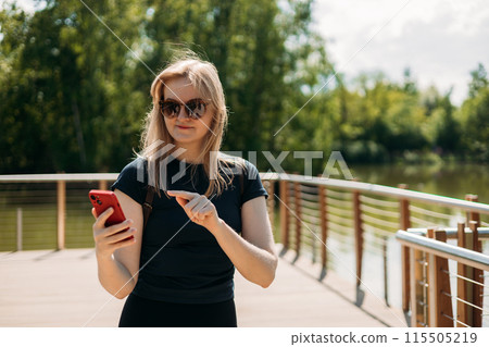 Happy blonde Woman in active sport clothes having a halt after hiking. Hiker with backpack holding smart phone near lake. Travel, technology and relax concept. 115505219