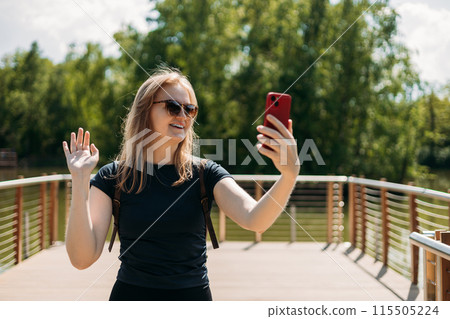 Active woman speaks via video link through the phone, takes a selfie near lake on nature background. Concept of an active lifestyle. Summer park 115505224