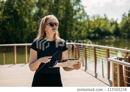 Stylish hipster woman in sunglasses holding paper map, wearing backpack and relaxing in nature. Travel and wanderlust concept. Amazing chill moment. Blonde 30s girl pointing finger on lake background. 115505236
