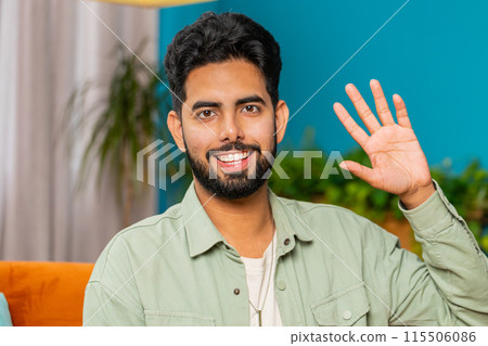 Indian man smiling friendly at camera, waving hands gesturing hello, hi, greeting at home on sofa 115506086