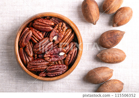 Pecan nuts in a wooden bowl on linen fabric. Dried pecan halves and whole pecans in shells, seeds and nuts of Carya illinoinensis. Ready to eat as snack, and also used for baking. Close-up from above. Pecan nuts in a wooden bowl on linen fabric. Dried pecan halves and whole pecans in shells, seeds and nuts of Carya illinoinensis. Ready to eat as snack, and also used for baking. Close-up from above. 115506352