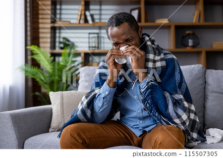 A man sneezing into a tissue, wrapped in a blanket, and sitting on a sofa. Depicts illness, cold, flu, and home comfort. 115507305