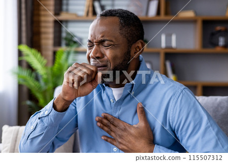 Man clutching chest and coughing, depicting symptoms of illness or respiratory distress in a home environment. 115507312