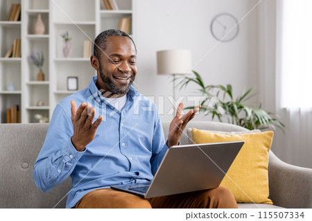 Smiling man having a joyful video call on his laptop while sitting on a cozy couch in his living room Smiling man having a joyful video call on his laptop while sitting on a cozy couch in his living room 115507334