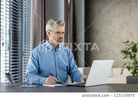 Senior man working on laptop in a modern home office. Focused and professional, he is taking notes on a notepad. Senior man working on laptop in a modern home office. Focused and professional, he is taking notes on a notepad. 115510973