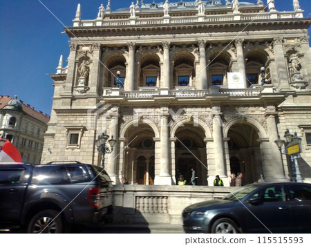 A beautiful building in an old style.There is a private vehicles parked along the fence, and behind there is a building with columns and statues. 115515593