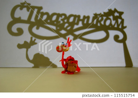 A toy dragon, its shadow and a shadow with an inscription. A little dragon and a congratulatory inscription Happy Holidays in Russian. A toy dragon, its shadow and a shadow with an inscription. A little dragon and a congratulatory inscription Happy Holidays in Russian. 115515754