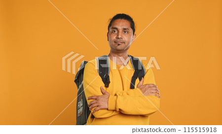 Portrait of confident indian man holding thermal backpack, crossing arms, studio background. Radiant nonchalant delivery person prepared to deliver takeaway food to customers, camera A Portrait of confident indian man holding thermal backpack, crossing arms, studio background. Radiant nonchalant delivery person prepared to deliver takeaway food to customers, camera A 115515918