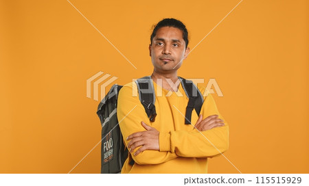 Portrait of upbeat indian man holding thermal backpack, isolated over studio background. Friendly cheerful person prepared to deliver takeaway food to customers, camera A 115515929