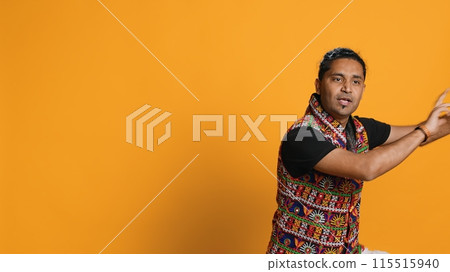 Ecstatic person wearing traditional indian attire doing energetic dance steps. Lively talented man doing entertaining bollywood choreography moves, studio background, camera A 115515940