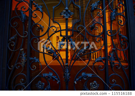 Beautiful, forged gate with many curls and leaves.Through the gate you can see the courtyard, illuminated by sunlight. Beautiful, forged gate with many curls and leaves.Through the gate you can see the courtyard, illuminated by sunlight. 115516188