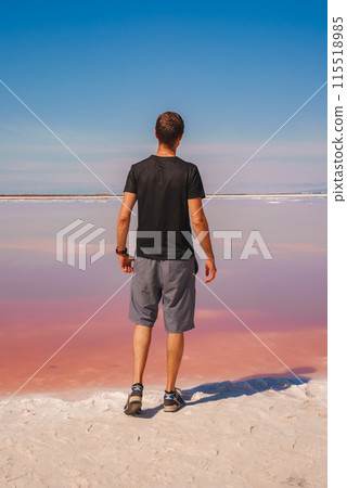 Man in black shirt and grey shorts stands at edge of pink lake in California. Scene showcases stunning contrast between pink water and blue sky at Alviso Pink Lake Park. Man in black shirt and grey shorts stands at edge of pink lake in California. Scene showcases stunning contrast between pink water and blue sky at Alviso Pink Lake Park. 115518985