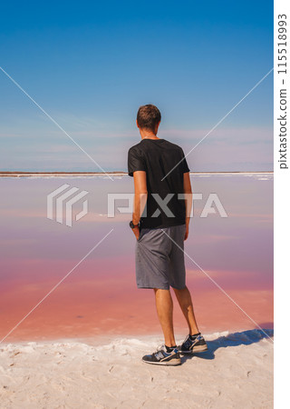Person in casual attire stands at edge of pink lake under clear blue sky at Alviso Pink Lake Park, CA. Unique coloration creates serene, surreal scene. 115518993