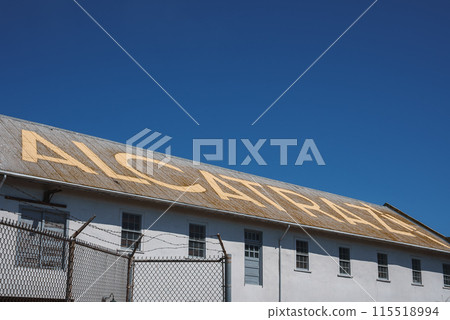 Close up view of white building at Alcatraz prison complex in San Francisco, USA. ALCATRAZ in bold yellow letters on roof, chain link fence with barbed wire in foreground. Sky is clear blue. 115518994