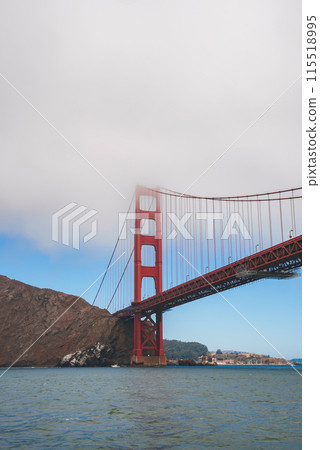 Iconic Golden Gate Bridge in San Francisco, CA. View from water level, showcasing bridge's famous orange color against blue sky. Tower dominates, cityscape in background. 115518995