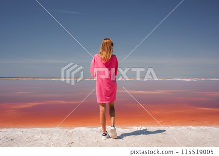 Person in pink dress stands at edge of vivid pink lake surrounded by white salt flats in Alviso Pink Lake Park, CA. Serene, surreal landscape with clear blue sky. 115519005