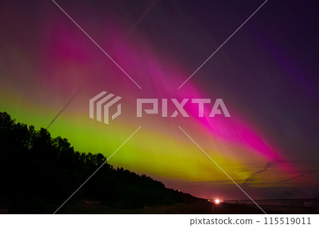 A stunning display of the aurora borealis illuminates the sky with green and pink hues over a coastal area near Riga and Jurmala, Latvia. A forest silhouette contrasts the vibrant colors. A stunning display of the aurora borealis illuminates the sky with green and pink hues over a coastal area near Riga and Jurmala, Latvia. A forest silhouette contrasts the vibrant colors. 115519011