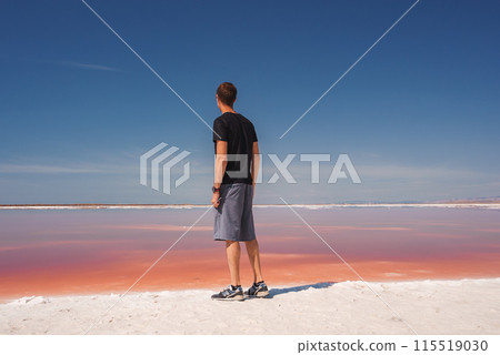 Person in black t shirt and grey shorts stands at pink lake in Alviso Pink Lake Park, California. Serene scene with striking colors and salt deposits. 115519030