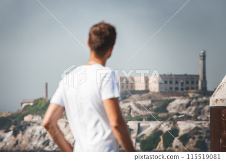 Person looking at Alcatraz Island from San Francisco, with historic prison visible in the distance. Clear weather and casual dress for a tourist like atmosphere. 115519081