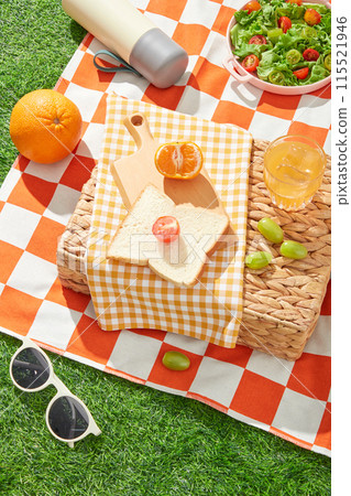 Creative template of picnic theme on grass, from top view. A yellow towel spread on a rattan basket with a wooden tray of sandwich and fruit above, below is an orange carpet contains picnic items 115521946