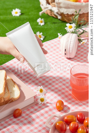 Cosmetics advertising photo with a picnic concept from a high angle, an empty label tube is held by a beautiful hand, below is a pink plaid rug containing some picnic items Cosmetics advertising photo with a picnic concept from a high angle, an empty label tube is held by a beautiful hand, below is a pink plaid rug containing some picnic items 115521947