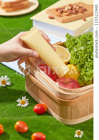 The unlabeled cosmetic tube in yellow color picked out of a wicker basket, which placed on green grass and containing some fresh vegetable and fruit for picnic meal. Photo of high angle, copy space The unlabeled cosmetic tube in yellow color picked out of a wicker basket, which placed on green grass and containing some fresh vegetable and fruit for picnic meal. Photo of high angle, copy space 115521948