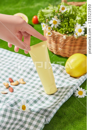 A yellow blank label cosmetic tube has just been placed on a blue carpet with a checkered pattern, behind is a green lawn, a rattan basket filled with feverfew flowers and a yellow lemon 115521950