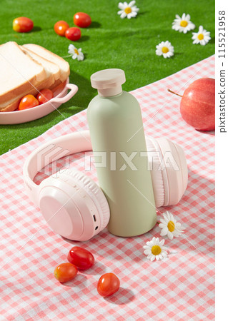High angle view photo on a green cosmetic bottle without label displayed on checkered pink picnic carpet, decorated by a headphone, some fruits and white feverfew flowers. Mock up for advertising 115521958