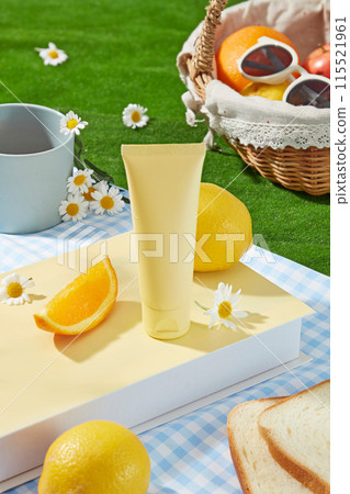 Outside picnic activity landscape, green grass containing a blue carpet with a yellow book, sandwich, citrus fruits and an empty label cosmetic tube in yellow color. High angle view, space for text 115521961