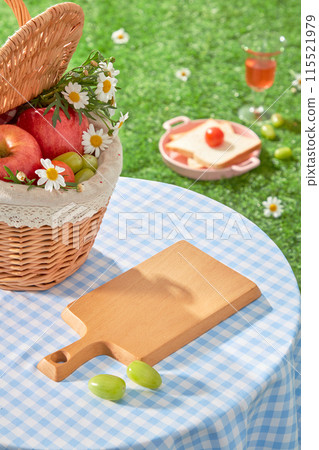 Outdoor picnic photo from above shot, a blank wooden tray placed on a checkered blue table amidst the grass, next to a rattan basket full of fruit. Vacant space for adding design elements 115521979
