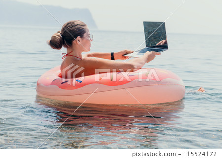 Woman freelancer works on laptop swimming in sea on pink inflatable ring. Happy tourist floating on inflatable donut and working on laptop computer in calm ocean. Freelance, remote working anywhere 115524172