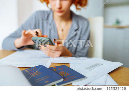 young woman, student, with red curly hair, fills out documents sitting at table at home, in foreground is passport citizen of Ukraine young woman, student, with red curly hair, fills out documents sitting at table at home, in foreground is passport citizen of Ukraine 115525426