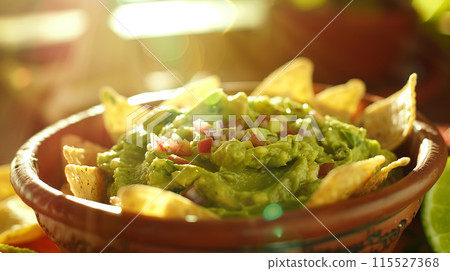 Freshly made guacamole, rich and green, with tortilla chips sprinkled around, shot in a sunny, appetizing setup. 115527368