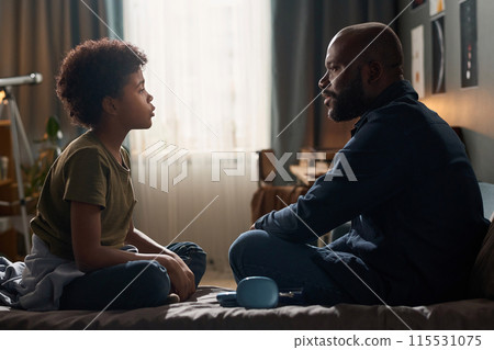 Side view portrait of African American family with single father and son talking to each other sitting on bed copy space 115531075