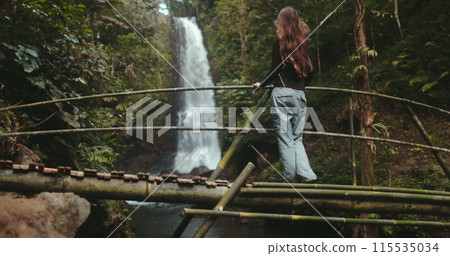 A woman stands confidently on a wooden bridge facing a majestic waterfall in Bali, Indonesia. Her presence adds scale to the powerful scene of water cascading downstream. A woman stands confidently on a wooden bridge facing a majestic waterfall in Bali, Indonesia. Her presence adds scale to the powerful scene of water cascading downstream. 115535034