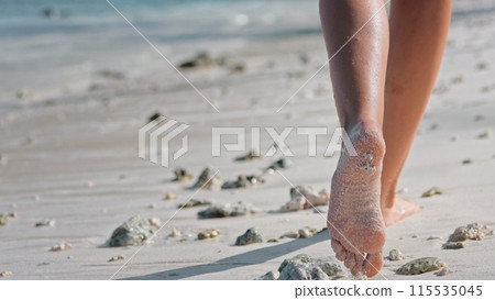 Person walking along sandy beach, feet leaving imprints on soft sand. Woman footsteps create a trail behind them as they stroll near the ocean waters edge, enjoying summer holiday vacation. Back view Person walking along sandy beach, feet leaving imprints on soft sand. Woman footsteps create a trail behind them as they stroll near the ocean waters edge, enjoying summer holiday vacation. Back view 115535045
